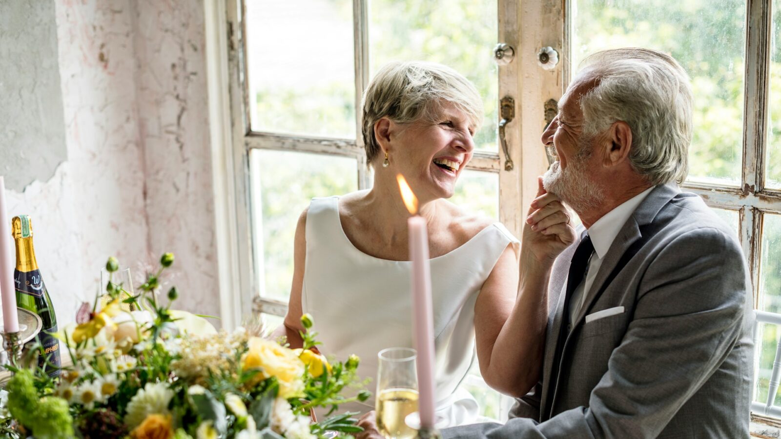 An older couple celebrates their wedding sitting behind a table covered in flowers, champagne, and tapered candles. The bride, with short gray hair and dressed in a wedding dress, smiles happily at the groom in a grey suit, while she lifts his bearded chin with her finger, representing second marriage contracts.