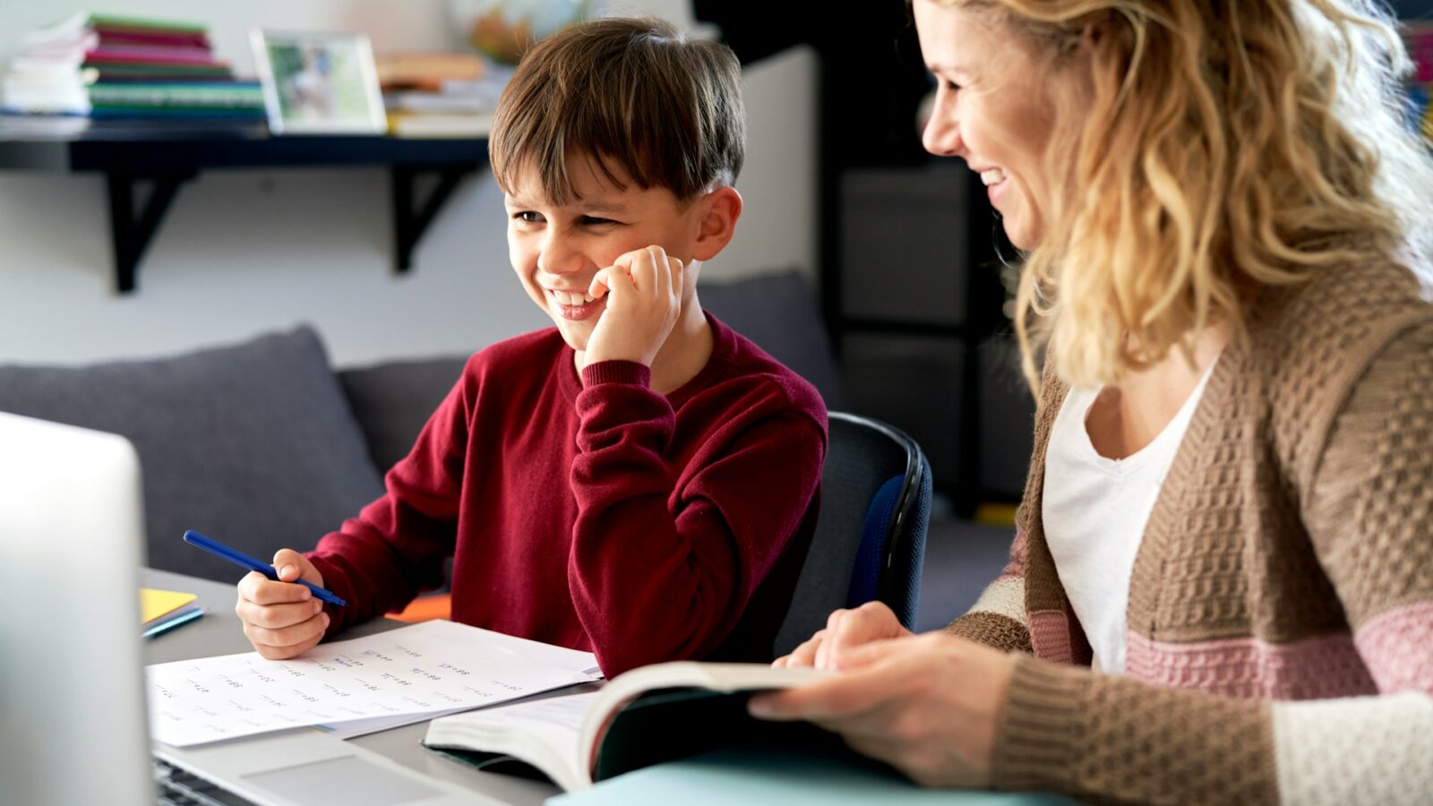 A young boy sits with his tutor at a desk in front of a laptop. He is filling out math problems on the papers in front of him, while she shows him information from the book open in front of her, representing tutoring expenses.