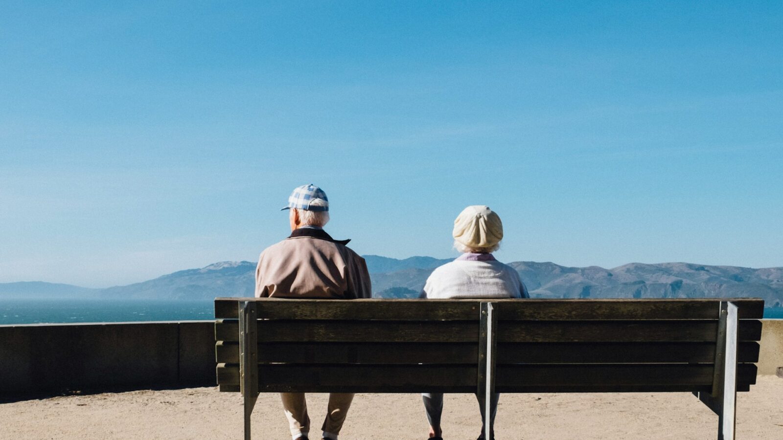 An elderly couple sits on a bench looking out over the sea and mountains with their backs to the camera, representing grey divorce.