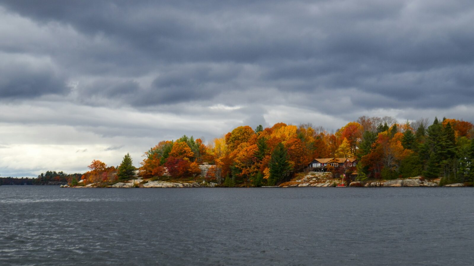 A gray cottage on a rocky point surrounded by autumn trees with dark clouds above and gray water stretched out before it, representing the family cottage during divorce.