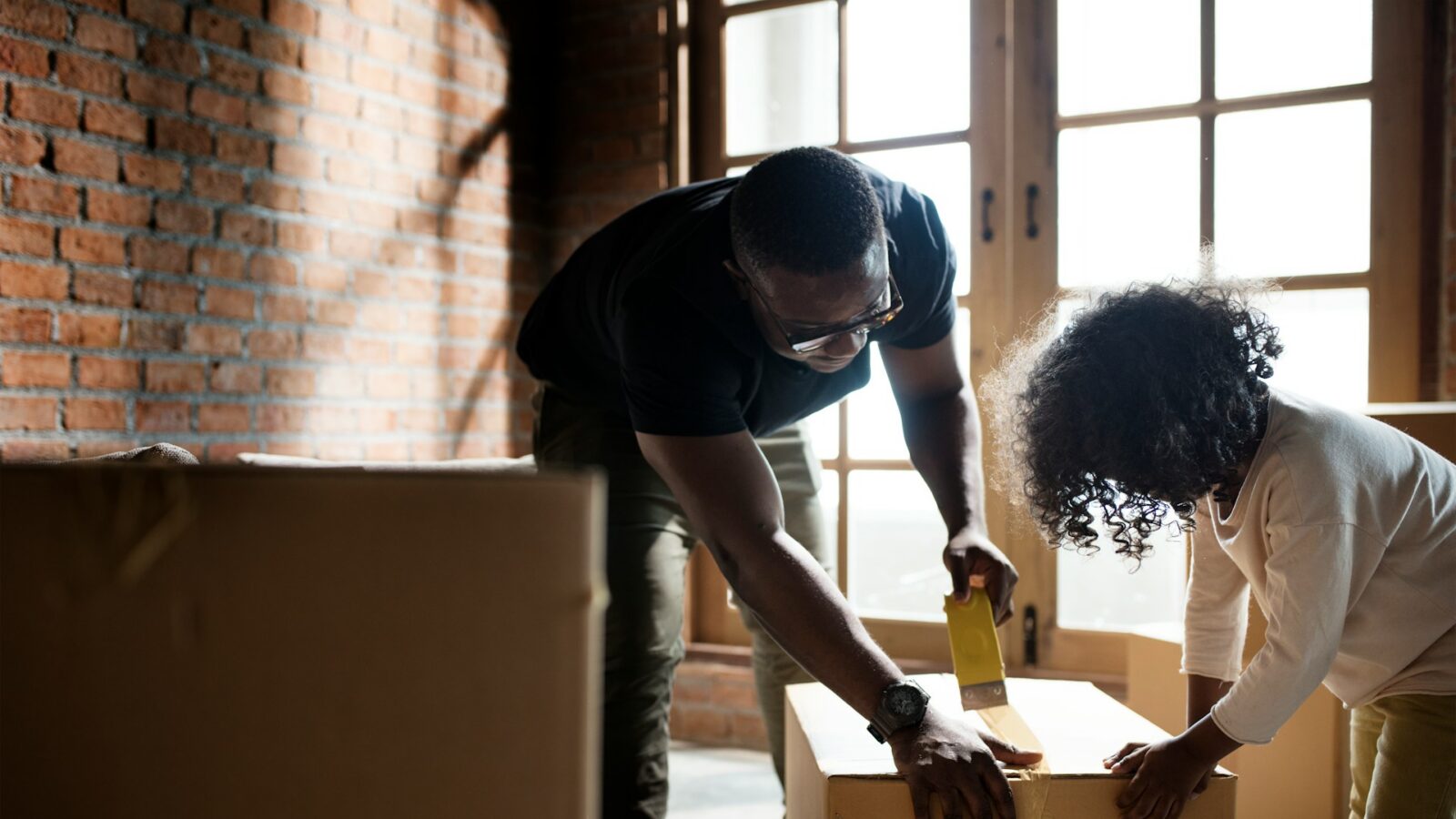 A man tapes closed a cardboard box with the help of his child, with another box off to the side, representing moving away with a child.