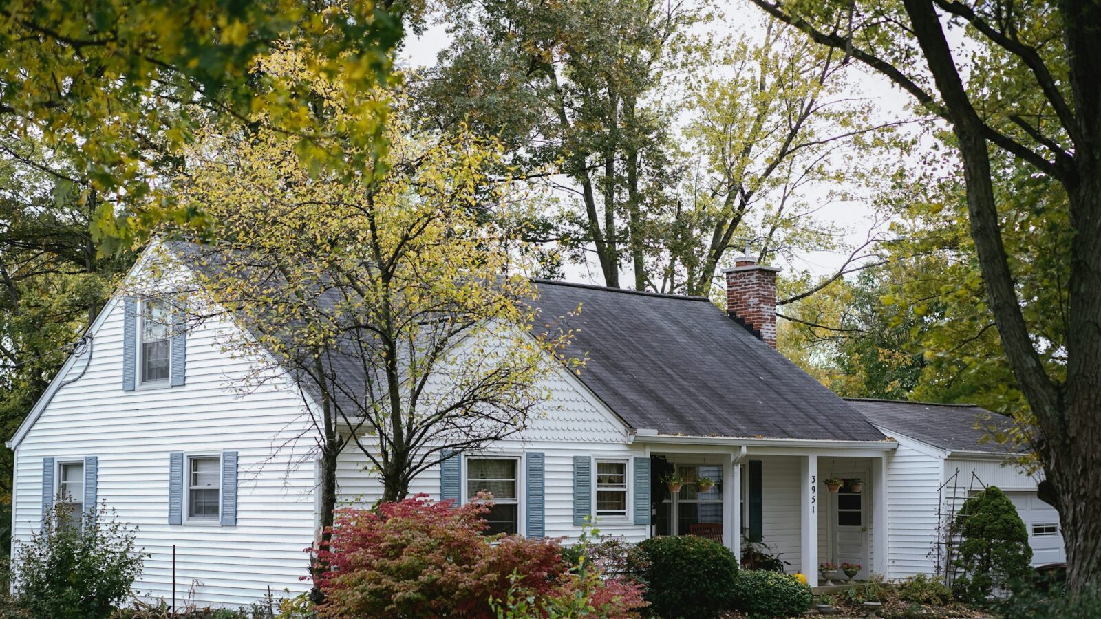A charming house with white sidind and blue shutters, a red brick chimney and an attached garage surrounded by green trees and shrubs,, representingproperty division during separation.