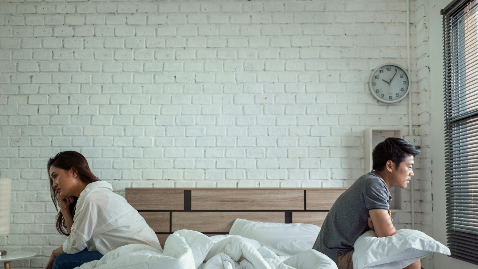 A husband and wife sit on opposite sides of an unmade bed with their backs to each other, looking dejected, representing what couples should do after a separation.