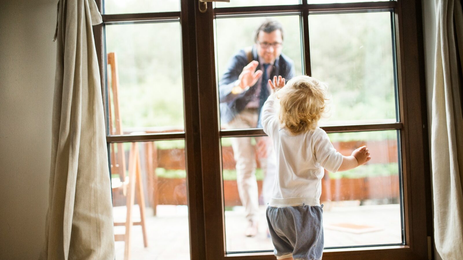 A toddler stands inside behind a set of French doors as his father approaches the door from outside smiling and waving, representing Parenting Plans.