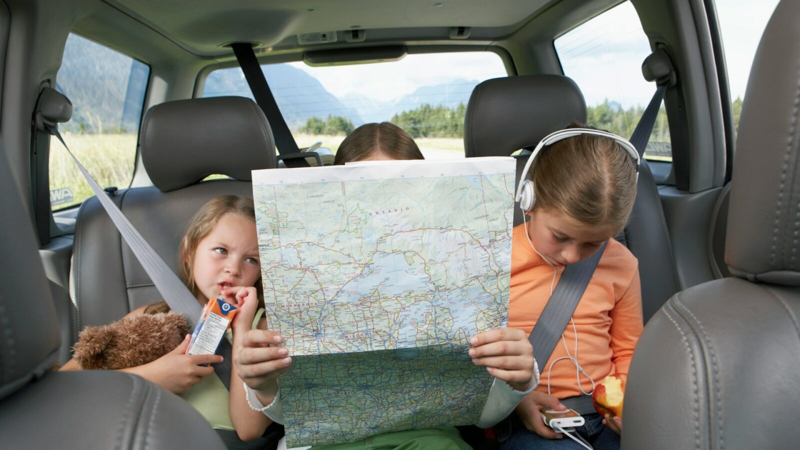Three siblings sit in the backseat of a car. The little girl on the left cubbles a stuffed bear and drinks from a juice box. The middle child his hidden by a map of Ontario, while th third eats an apple and lisyens to music on her headphones, representing taking children outside Ontario for summer vacation.
